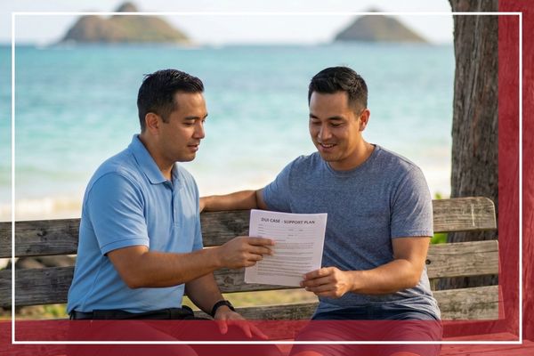 Two men sitting outdoors near a coastline reviewing and signing bail release documents.